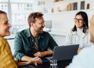 Vier Personen sitzen gemeinsam an einem Konferenztisch, lachen und unterhalten sich. Eine Person arbeitet an einem Laptop. Die Atmosphäre wirkt freundlich und kollegial.