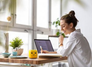 Junge Frau sitzt an einem modernen Schreibtisch mit Laptop, Notizbuch und einem gelben Würfel mit Gesicht, während sie lächelnd schreibt. Im Hintergrund Pflanzen und Tageslicht durch große Fenster.